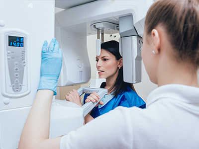 A woman in a blue lab coat stands beside a large, white 3D scanner, with a smiling technician wearing gloves and a face mask.