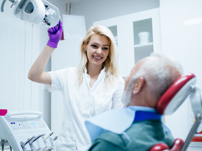 A woman in a white coat stands over an elderly man sitting in a dental chair, holding a mirror and smiling.