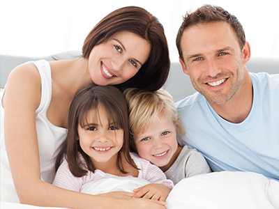 A family of four, including a man, woman, and two young children, smiling together in a cozy bedroom setting.