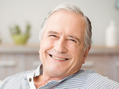 The image shows an older man with grey hair, wearing a blue shirt, smiling and looking directly at the camera. He is seated in a relaxed pose.