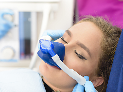 The image shows a person lying down with their eyes closed, wearing an oxygen mask and having their face covered by a blue device connected to the mask. A medical professional is attaching the device to the mask.