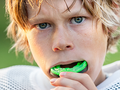 A young boy with blonde hair and blue eyes, wearing a white football jersey, brushing his teeth with a green toothbrush.