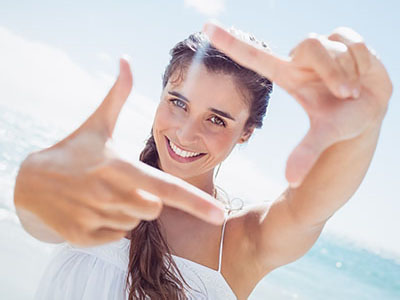 A young woman with long hair is smiling and holding up a peace sign against a bright, clear sky.