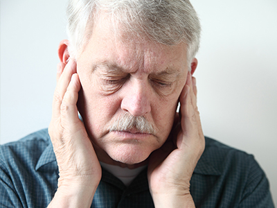 Man with gray hair, wearing a blue shirt and glasses, holding his hand to his ear in a state of distress or discomfort.