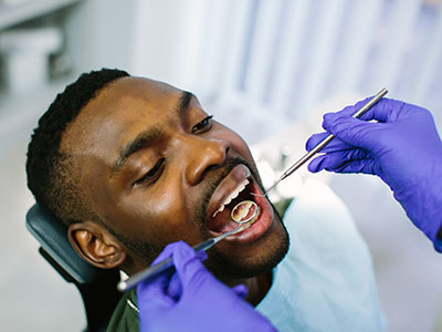 The image shows a person receiving dental care, with a dentist performing a procedure on their teeth while wearing gloves and using dental instruments.